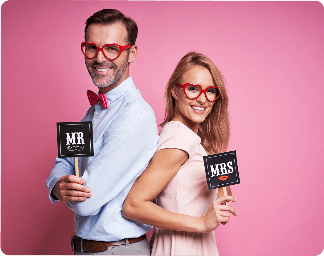A man and woman in glasses holding photo booth signs against a pink background.