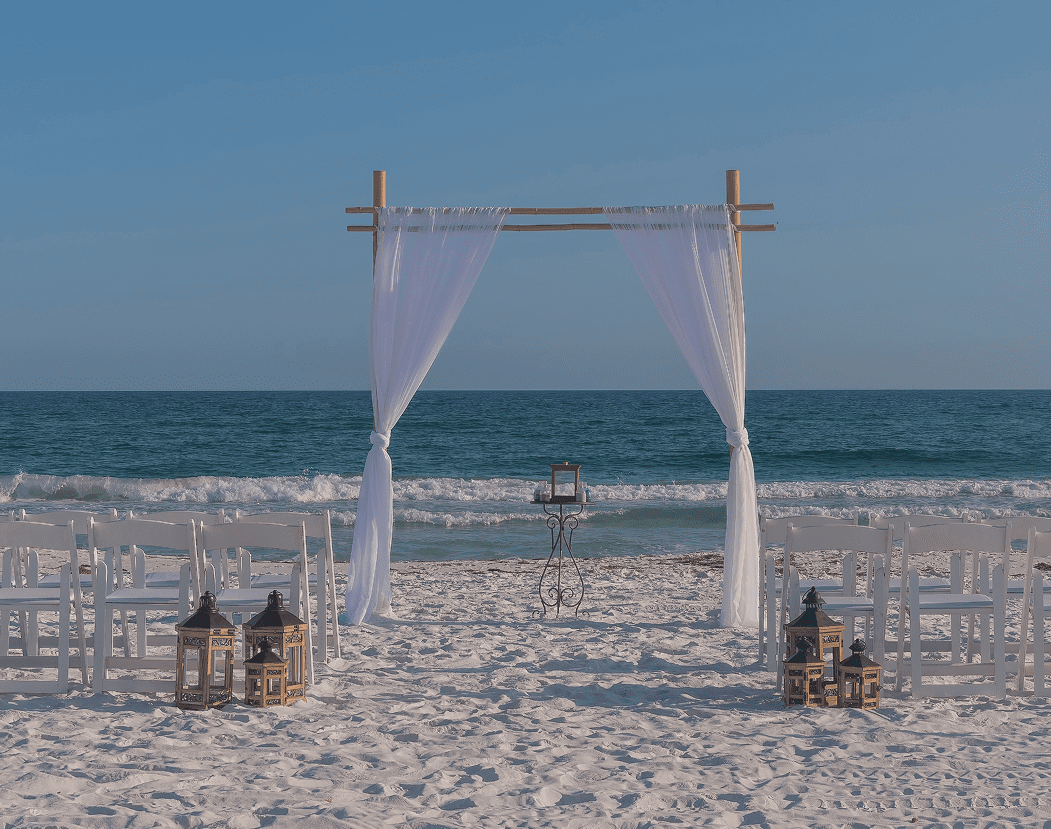 Elegant beach wedding arch with white drapes and lanterns at sunset.