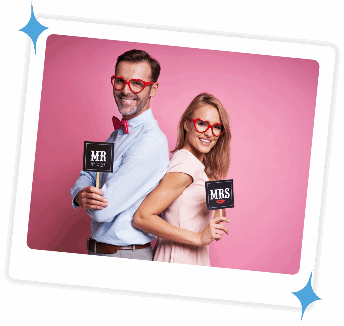A man and woman in glasses holding photo booth signs against a pink background.